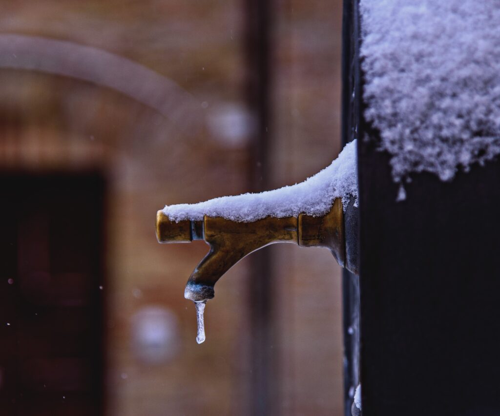 A close-up on an outdoor faucet, dusted with snow, with a frozen drip coming out of it—illustrating the importance of winterizing outdoor faucets to prevent frozen pipes.