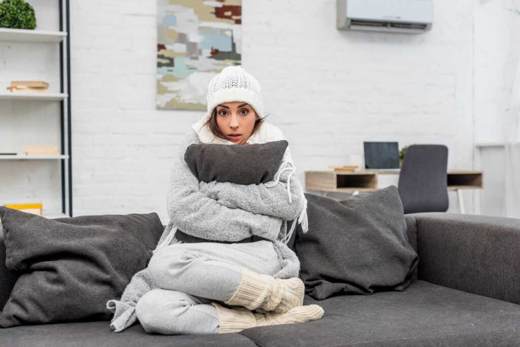 Cold young woman in layers of warm clothing sitting in home, hugging a cushion.