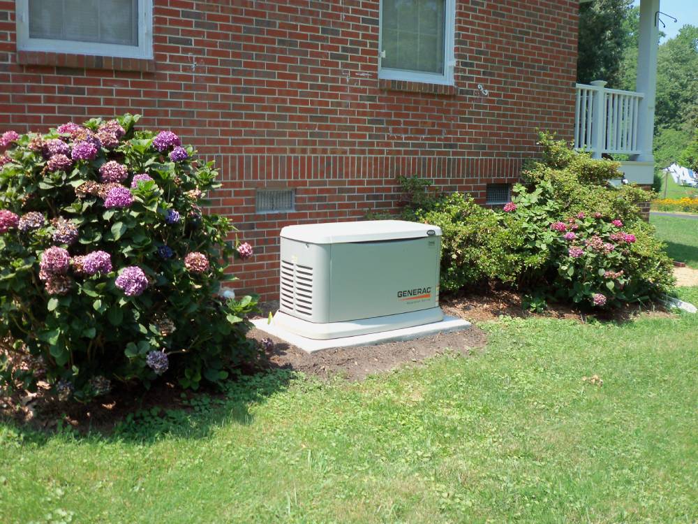A whole home generator sits next to a house with bushes on either side.