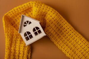Small wooden house with a bright yellow knit scarf wrapped around it, symbolizing the cozy warmth that can come from smart grades for your home heating system.