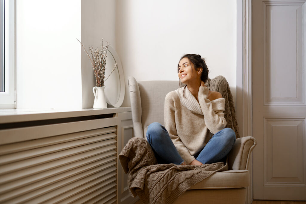A brunette woman in a beige sweater sits in a bright room, enjoying the smart thermostat benefits of efficient home heating and cooling.